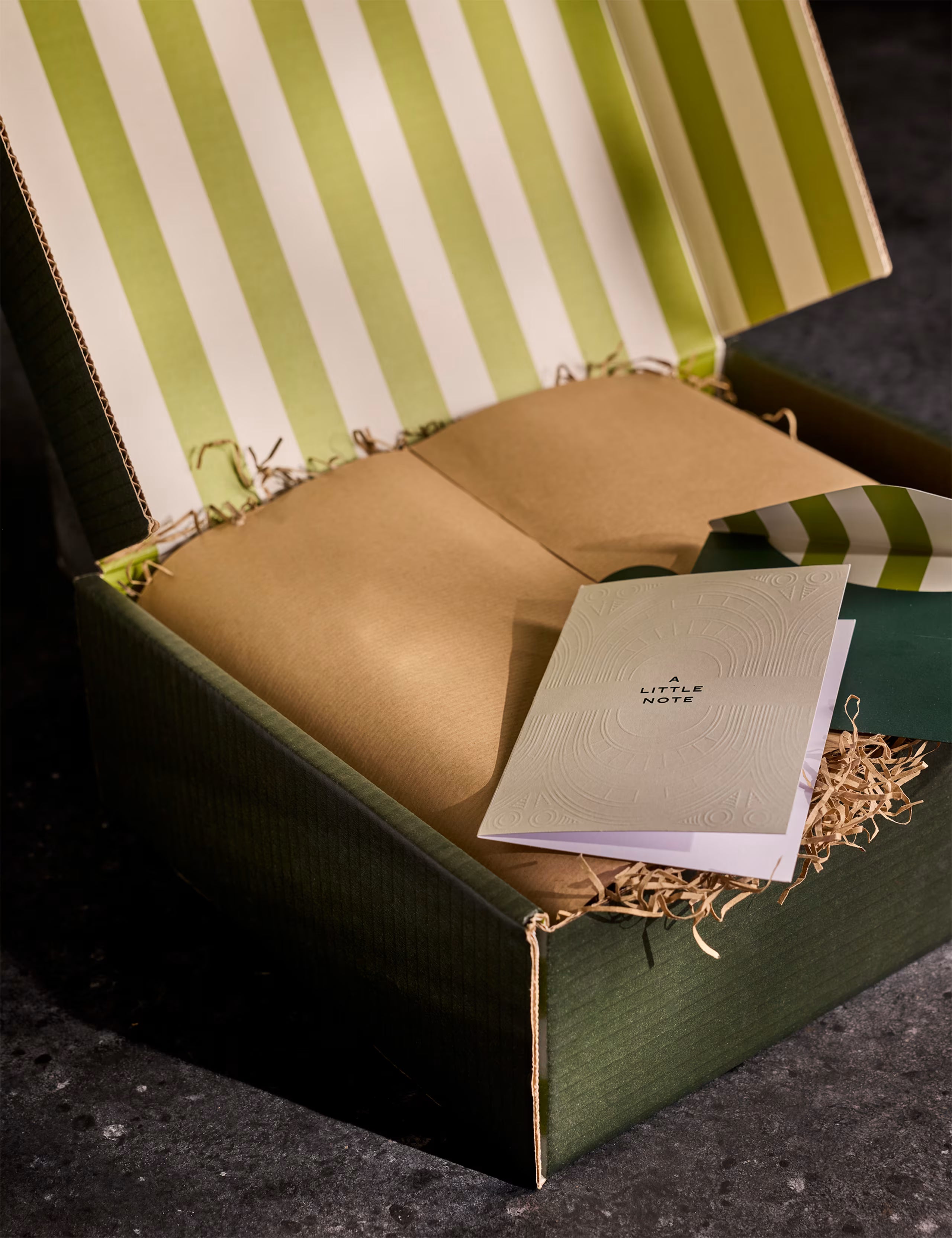 Open box with a notebook titled 'Little Note' and a brown envelope inside, against a striped background.