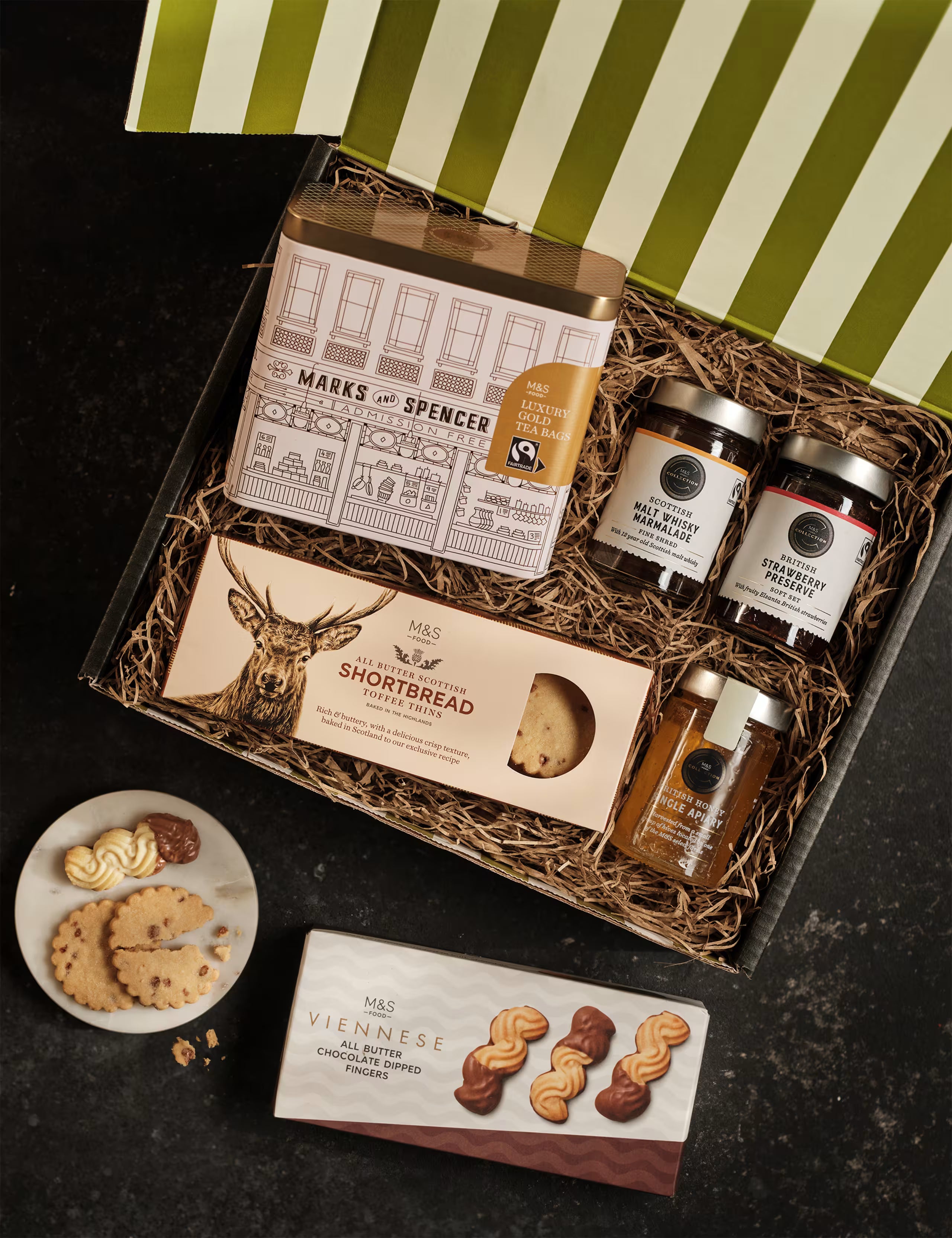 Gift basket with M&S products including jars, a box of shortbread, and cookies on a dark surface.